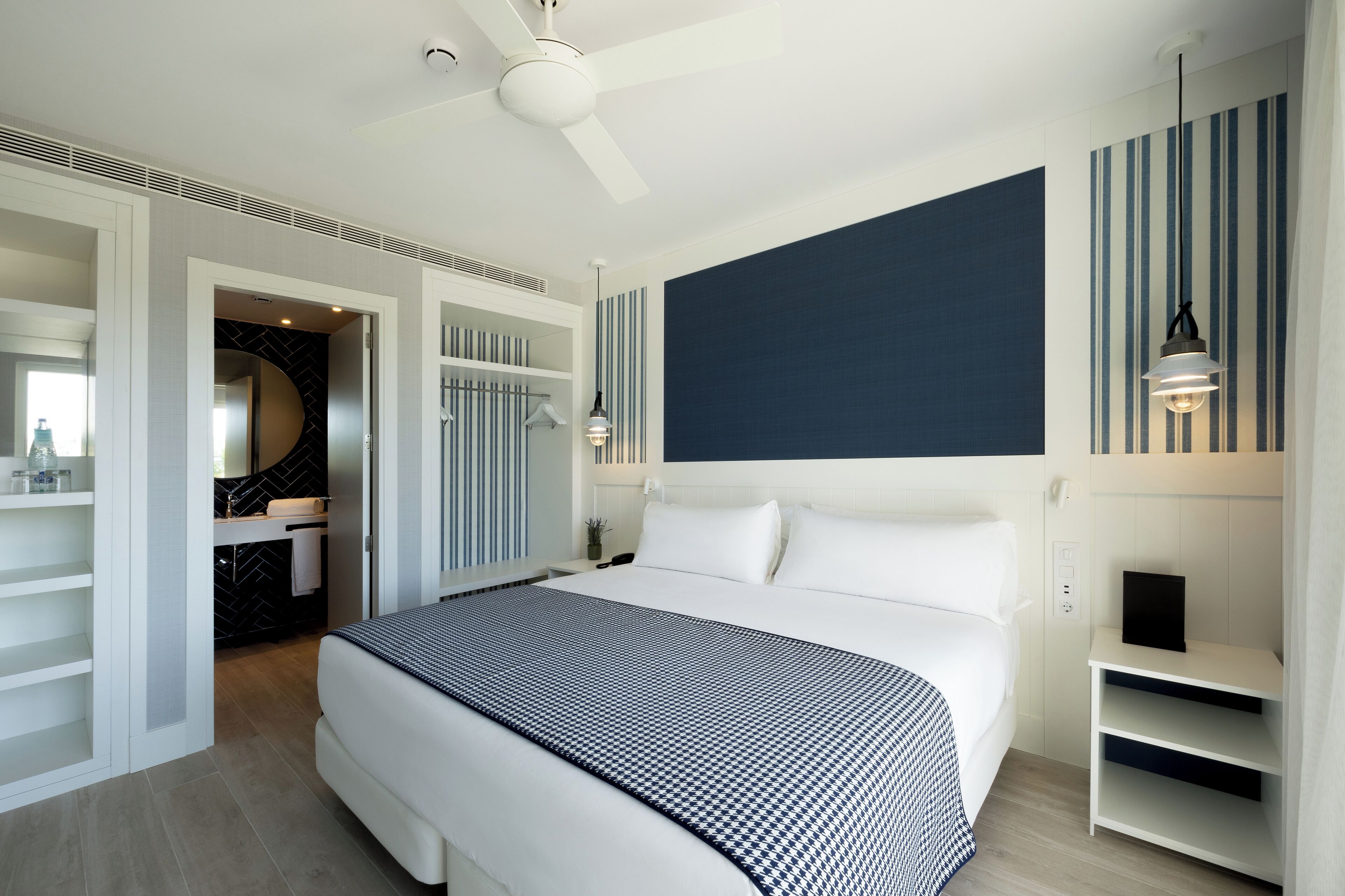 A modern hotel room with a large bed, a walk-in closet, a bathroom with a black and white tiled backsplash, and a dark accent wall.