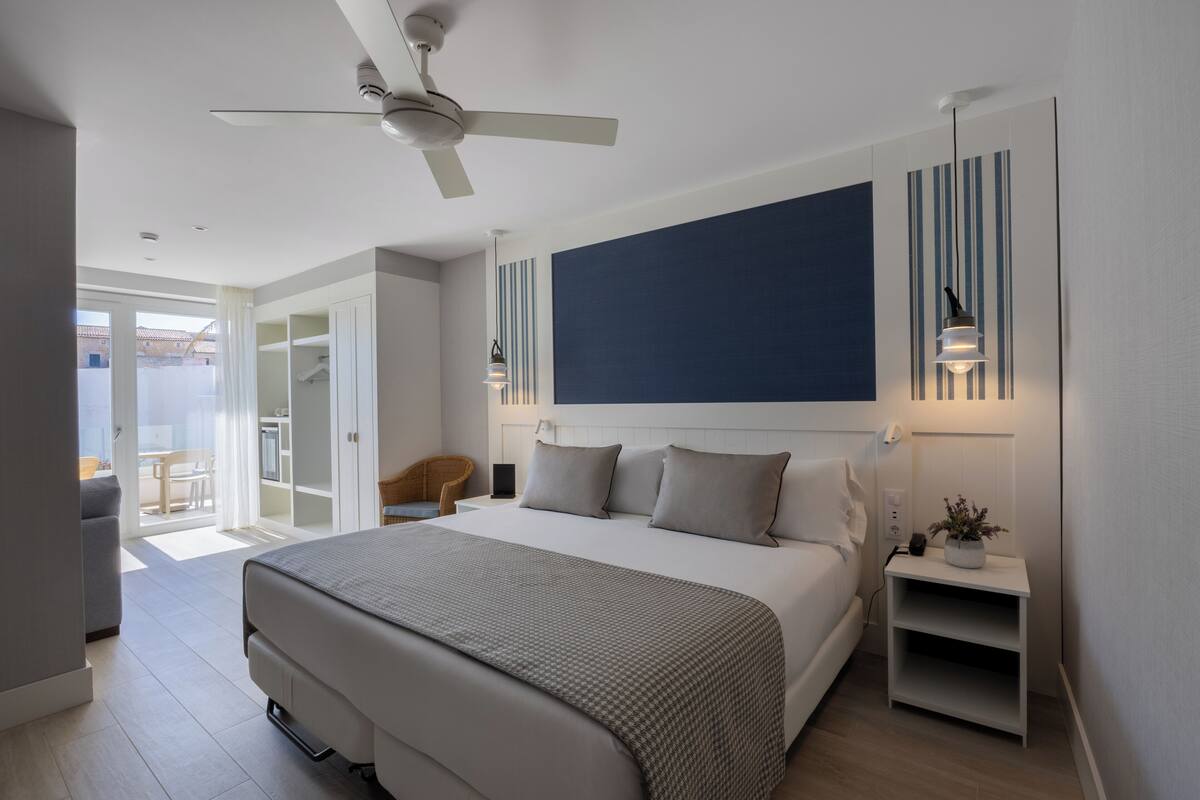 A modern hotel room with a large bed, a ceiling fan, a bedside table with a lamp, and a view of the outdoor area.