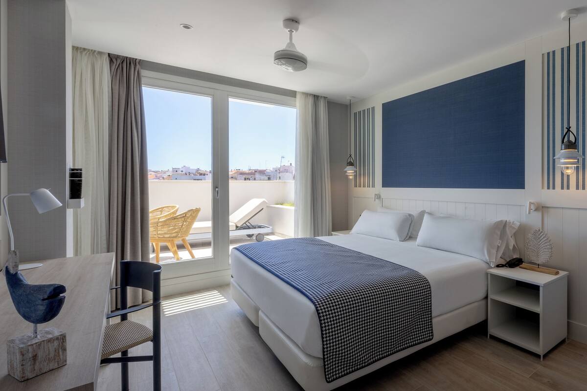 A modern hotel room with a large bed, a desk, and a chair. There is a balcony with a view of the city.