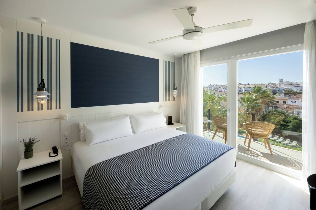 A modern hotel room with a large bed, a ceiling fan, a balcony with a view of the city, and a small table with a plant.