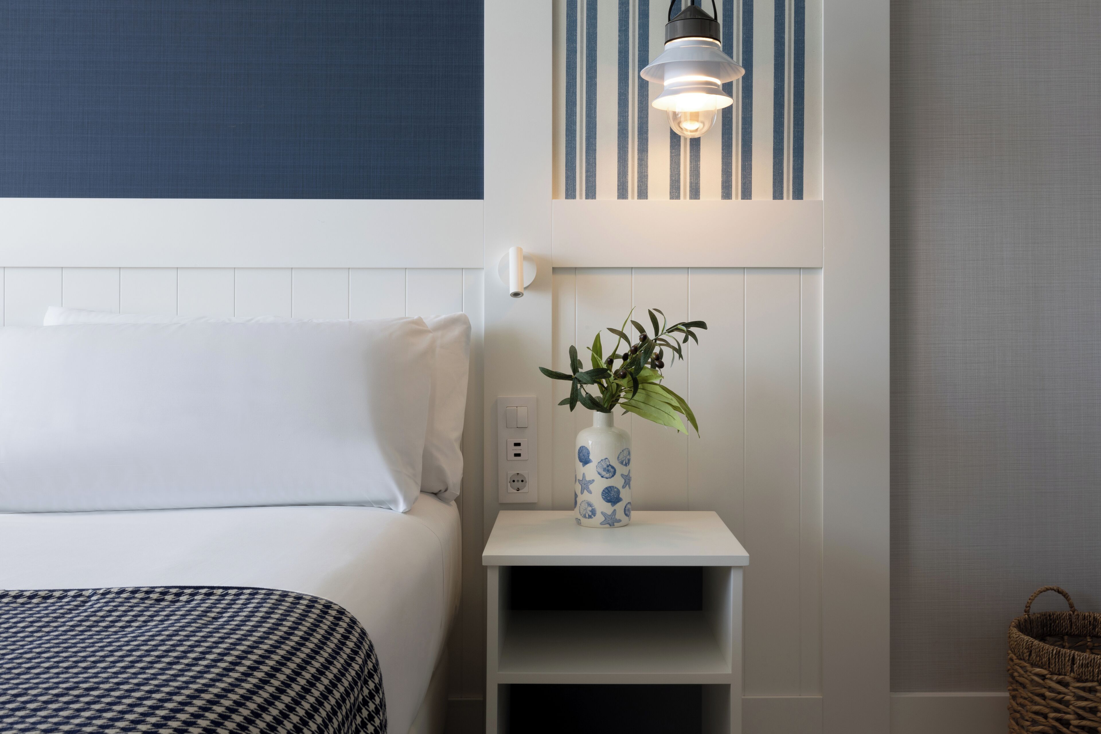 Une chambre d’hôtel avec un lit, une table de chevet ornée d’un vase de fleurs, une lampe fixée au mur et un panier posé sur le sol.