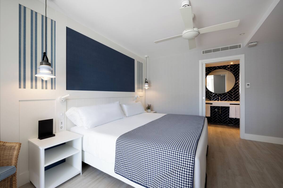 A modern hotel room with a large bed, a bedside table with a lamp, a ceiling fan, and a view of the bathroom.