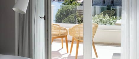 Et moderne hotelværelse med en seng, en hvid lampe og en balkon med fletstole samt udsigt til en historisk bygning og grønne områder.