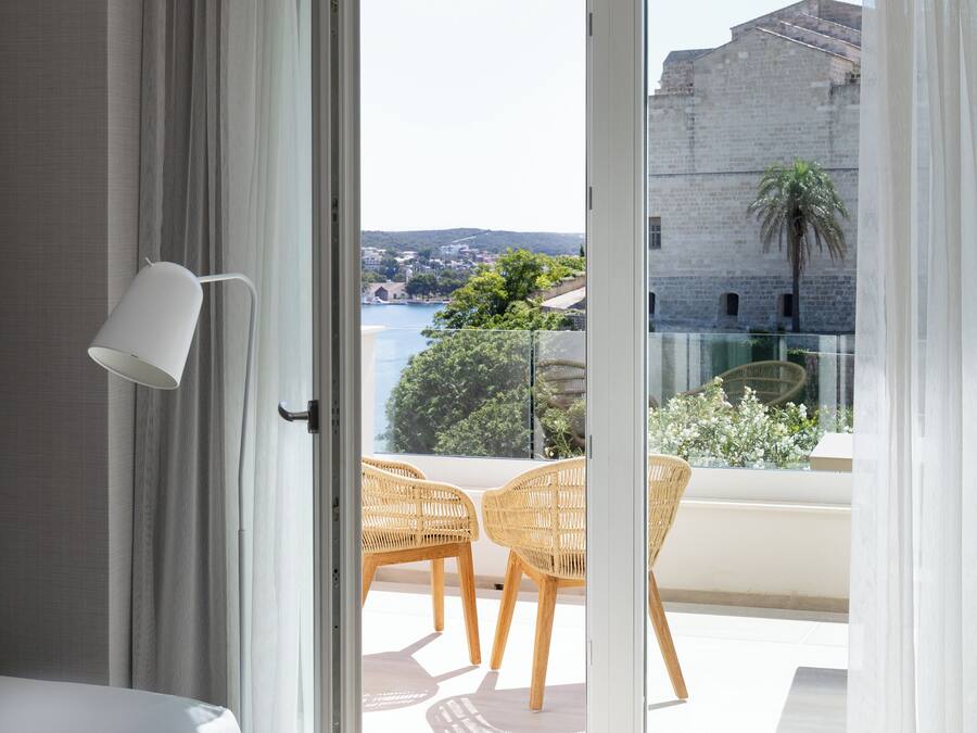 Una habitación de hotel moderna con cama, una lámpara blanca y un balcón con sillones de mimbre y vistas a un edificio histórico y vegetación.