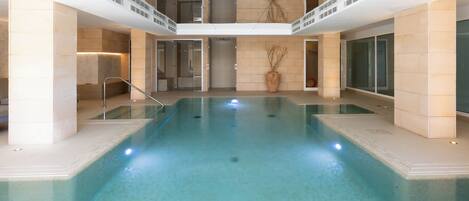 An indoor swimming pool with clear water, surrounded by stone walls and columns, and a second level balcony with railings.