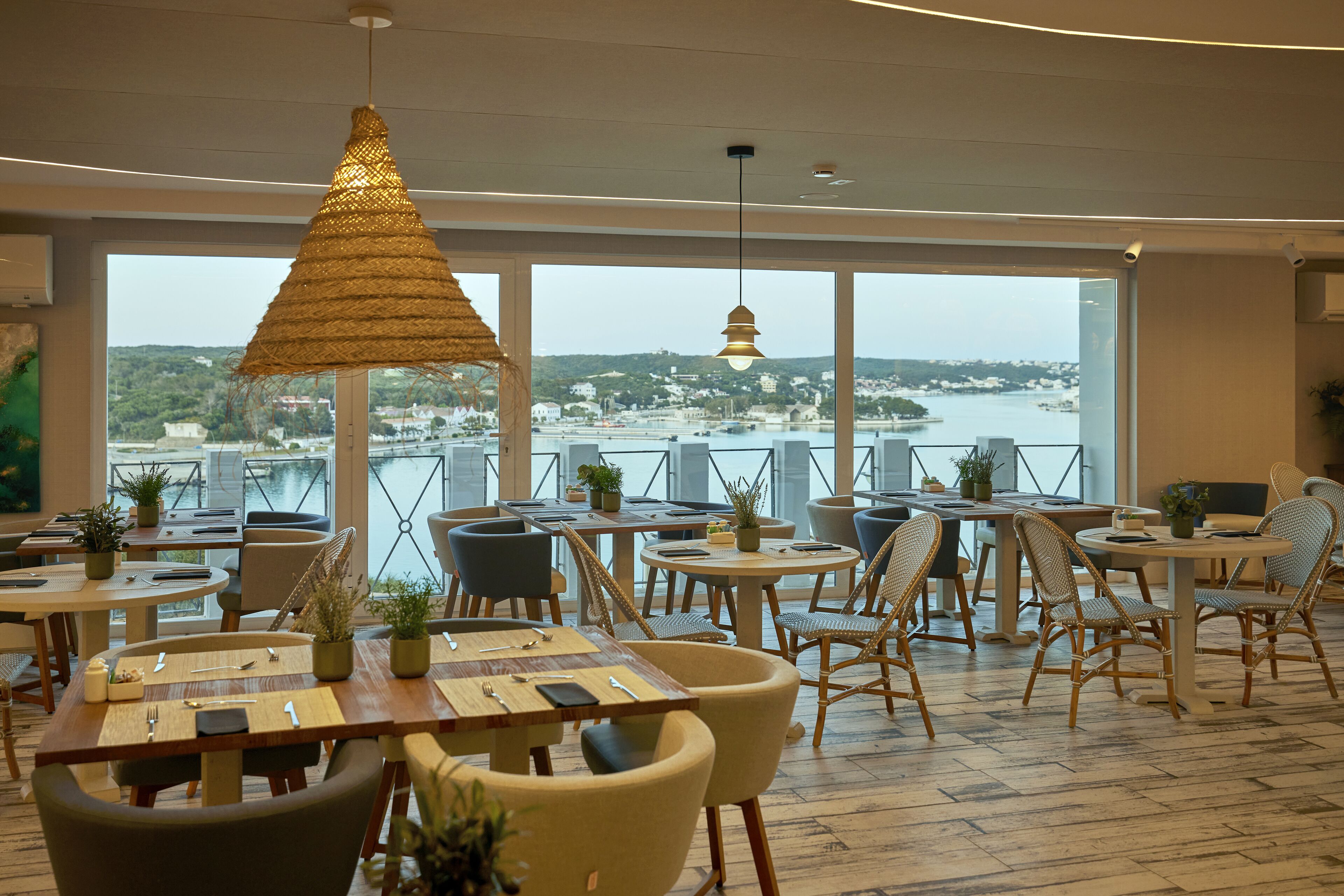 A restaurant with tables and chairs arranged for dining, large windows offering a view of a body of water and a distant town.