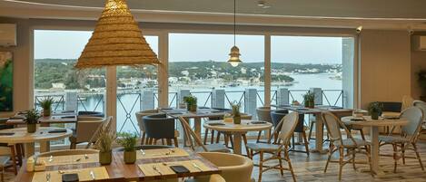 A restaurant with tables and chairs arranged for dining, large windows offering a view of a body of water and a distant town.
