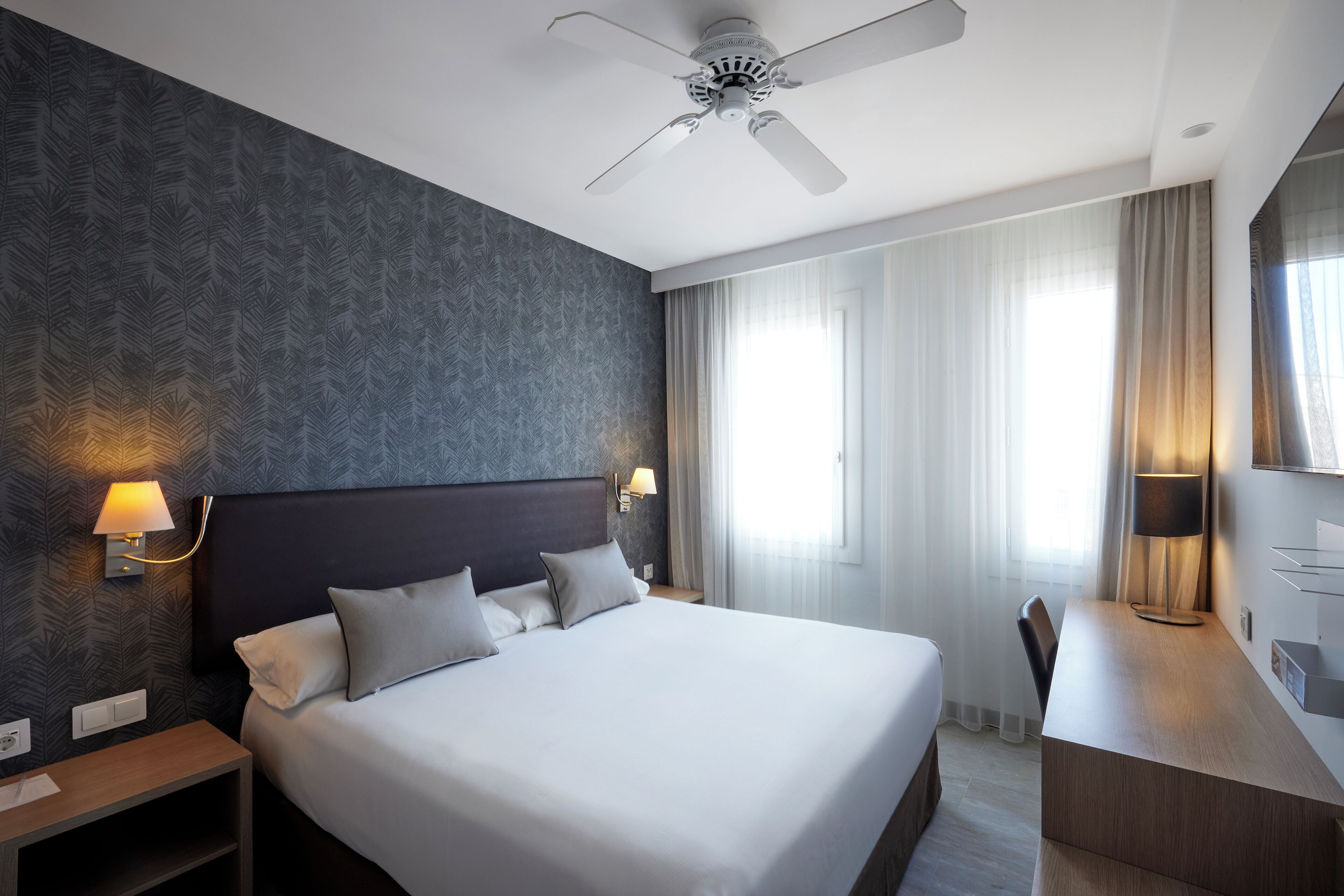 A modern hotel room with a large bed, a desk, a ceiling fan, and a window with curtains.