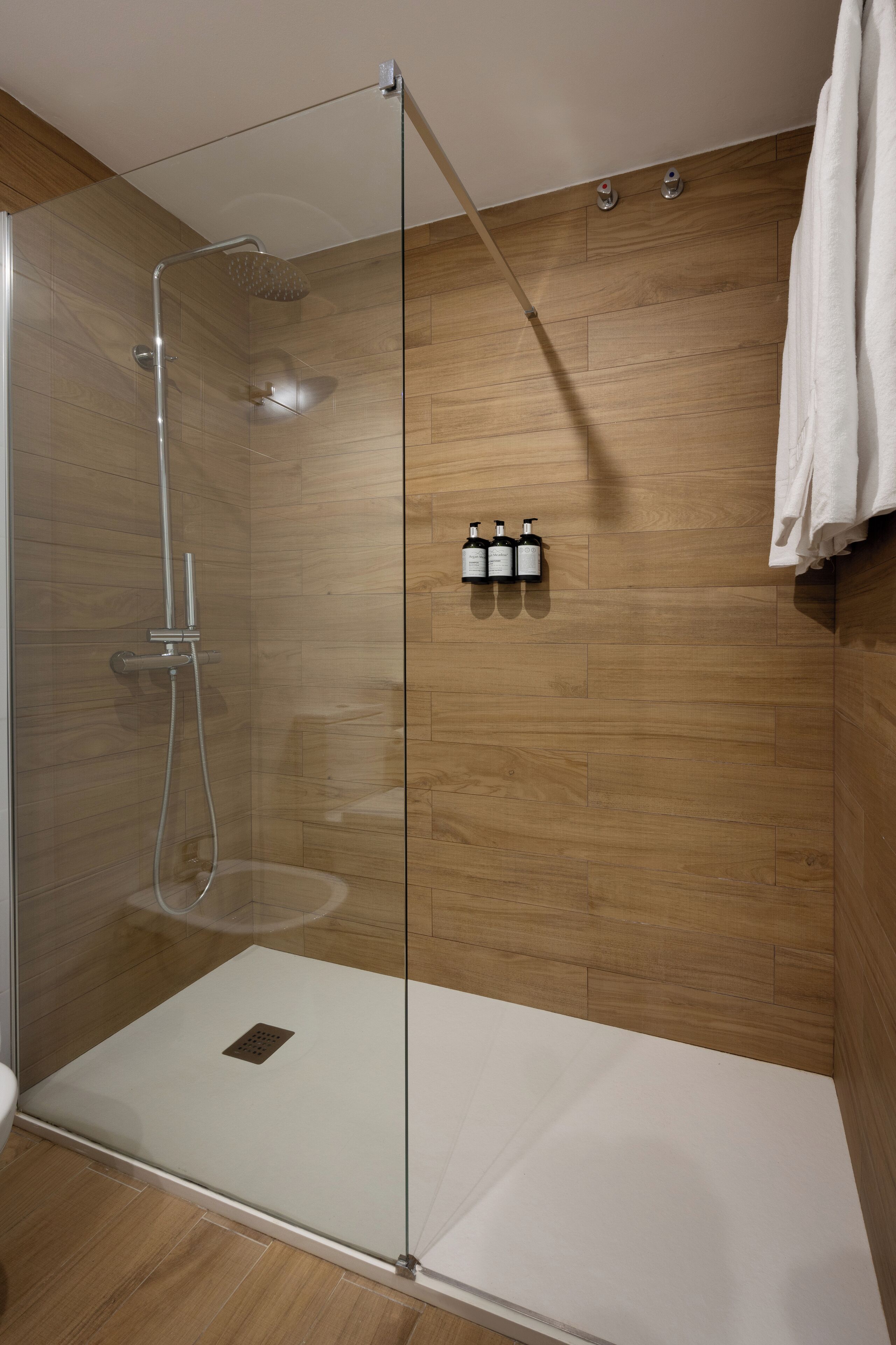 A glass-enclosed shower with a wooden floor, a towel hanging on the wall, and three bottles of shampoo.