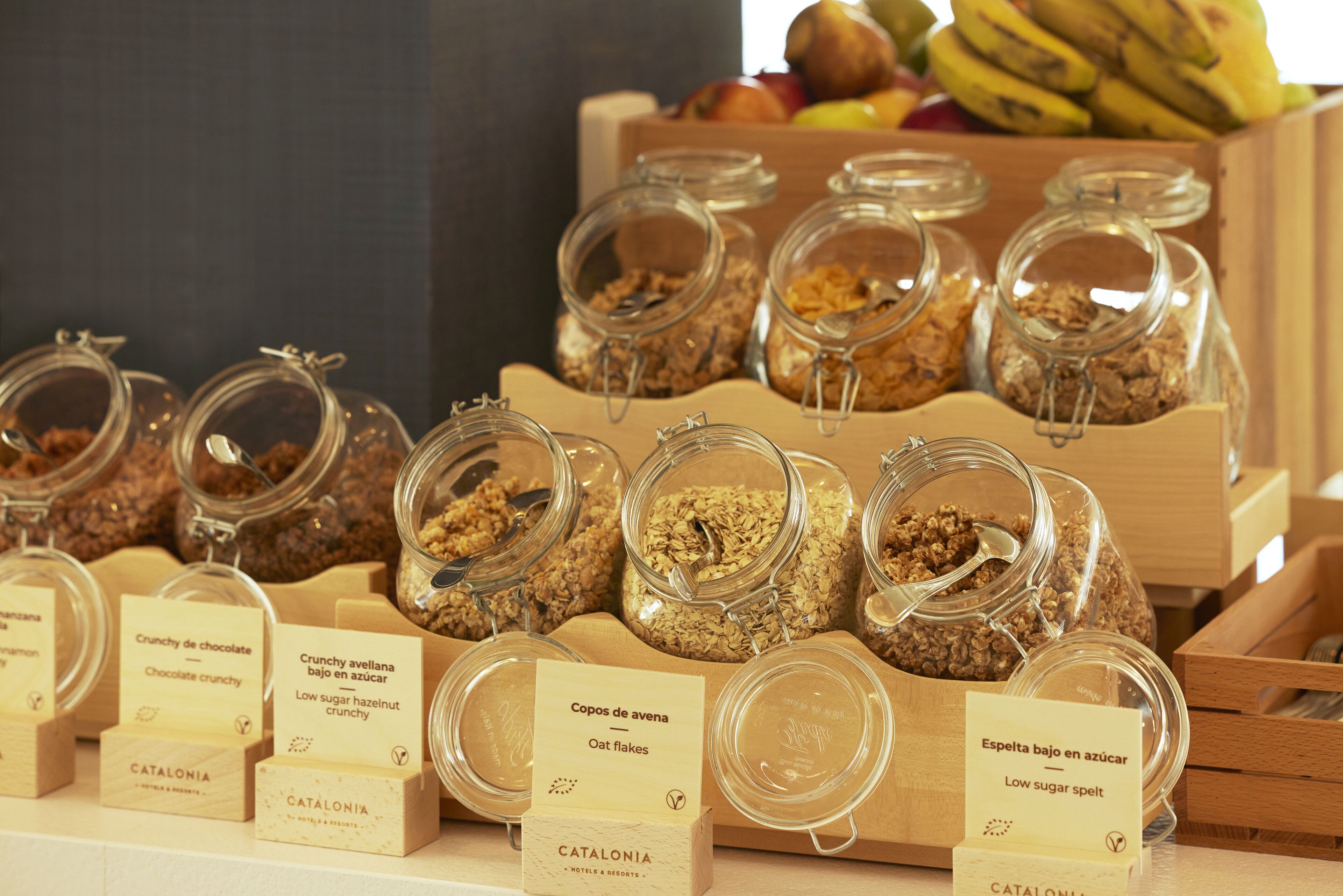 A display of various food items in glass jars with labels, including 'Crunchy de chocolate' and 'Oat flakes', arranged on a wooden table.