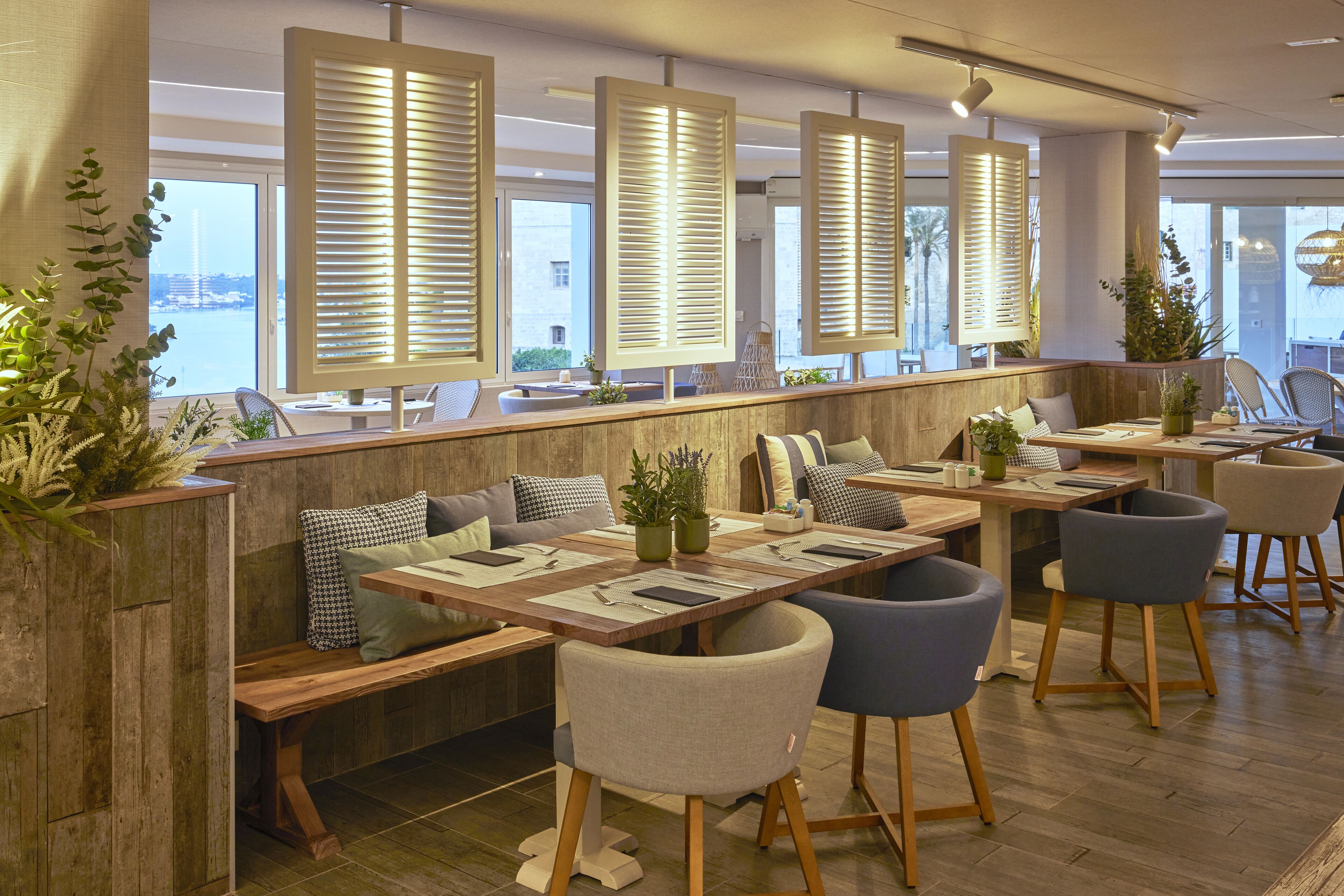 A modern restaurant with wooden tables, cushioned seating, and large windows with blinds.