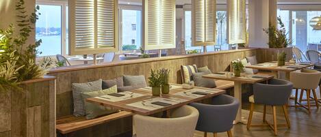 A modern restaurant with wooden tables, cushioned seating, and large windows with blinds.