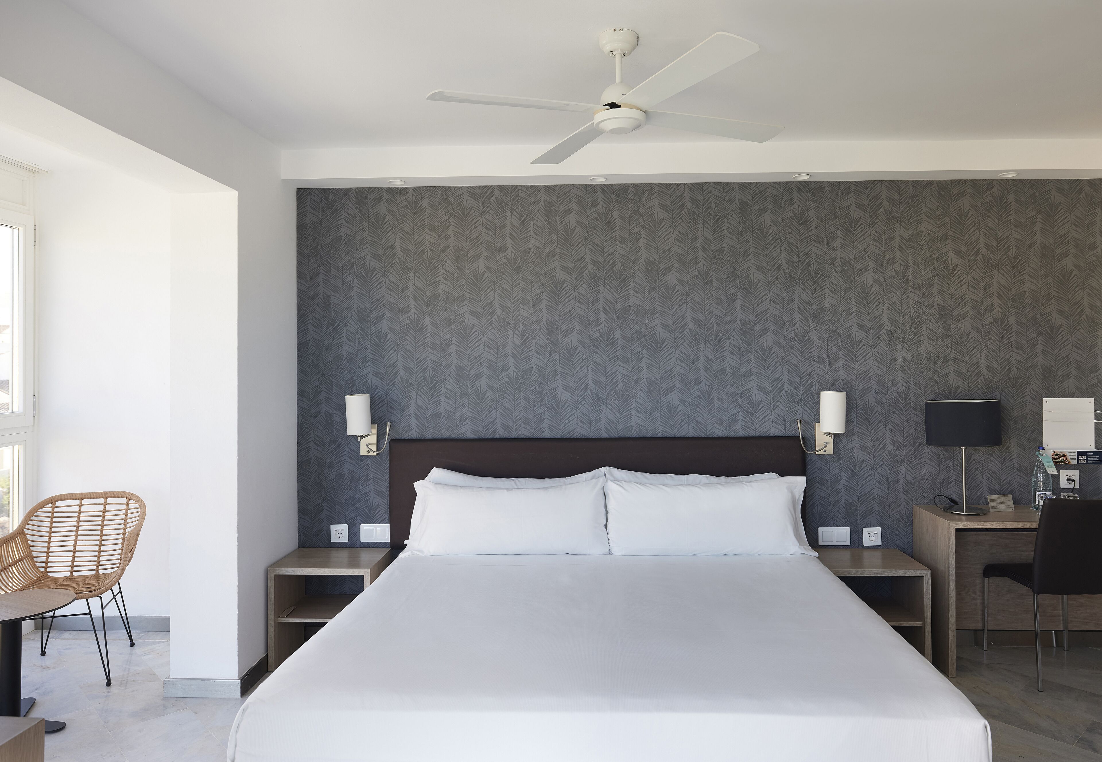 Habitación de hotel con una cama grande, ventilador de techo, escritorio con silla y lámpara de pared.