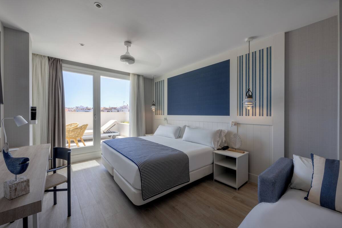 A modern hotel room with a large bed, a desk, and a chair. There is a view of the city through the window.