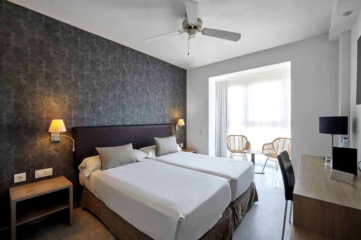A hotel room with two beds, a desk, a chair, a ceiling fan, and a window with curtains.