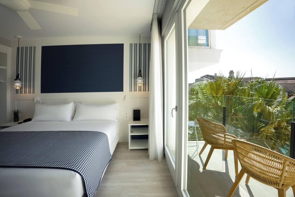 A modern bedroom with a large bed, a bedside table, a nightstand, a wall-mounted TV, and a balcony with wicker chairs and a view of palm trees.