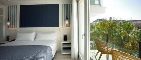 A modern bedroom with a large bed, a bedside table, a nightstand, a wall-mounted TV, and a balcony with wicker chairs and a view of palm trees.