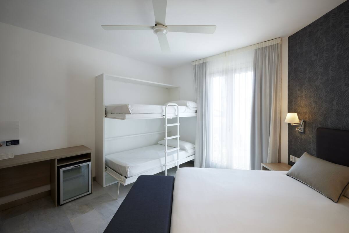A hotel room with a bunk bed, a desk with a refrigerator, a bedside lamp, and a ceiling fan.