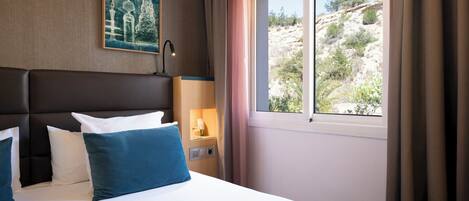 Una habitación de hotel con cama, una mesita de noche con lámpara, un cuadro en la pared y una ventana con cortinas.