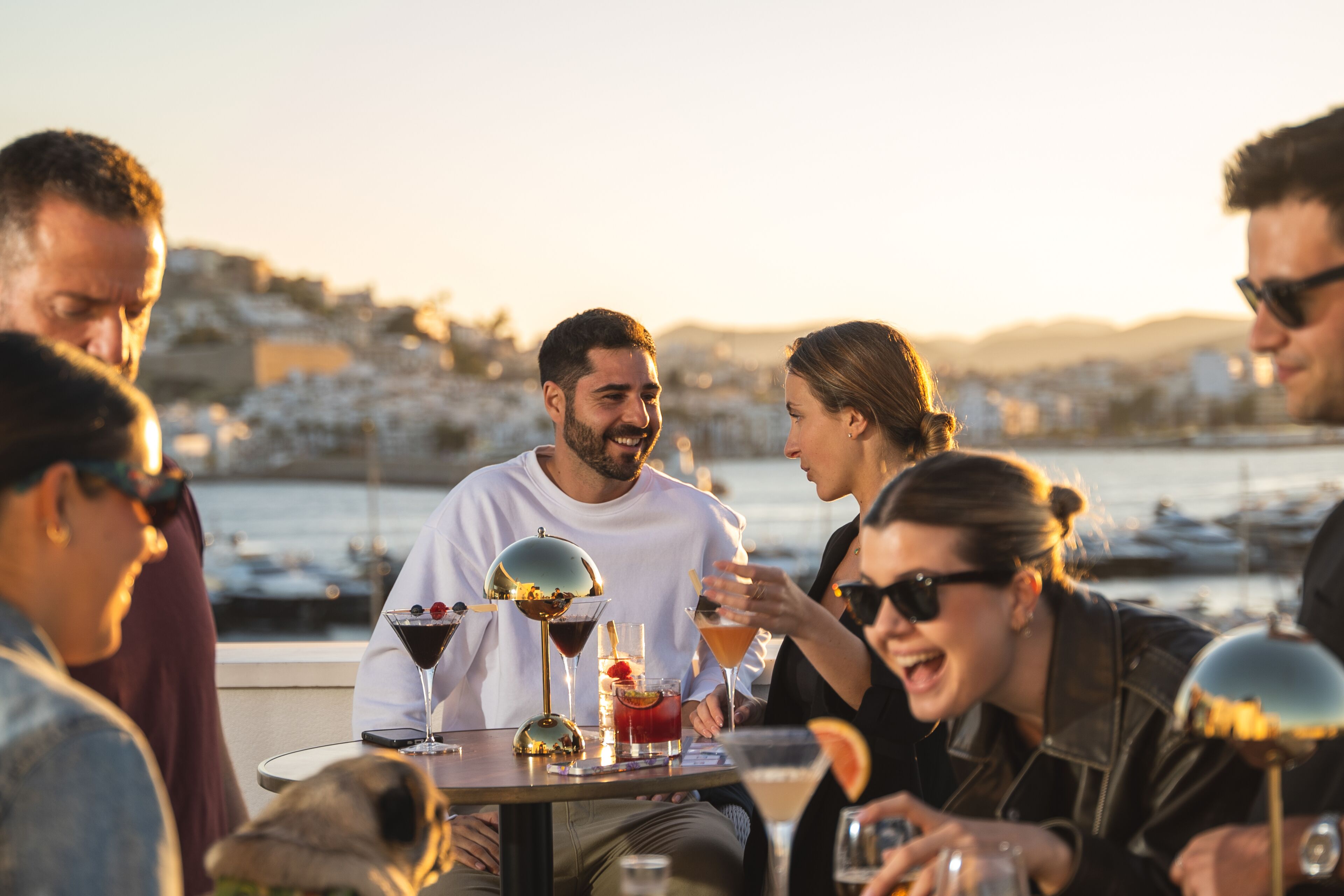 A group of people sitting at a table with drinks, overlooking a marina with boats and buildings in the background.