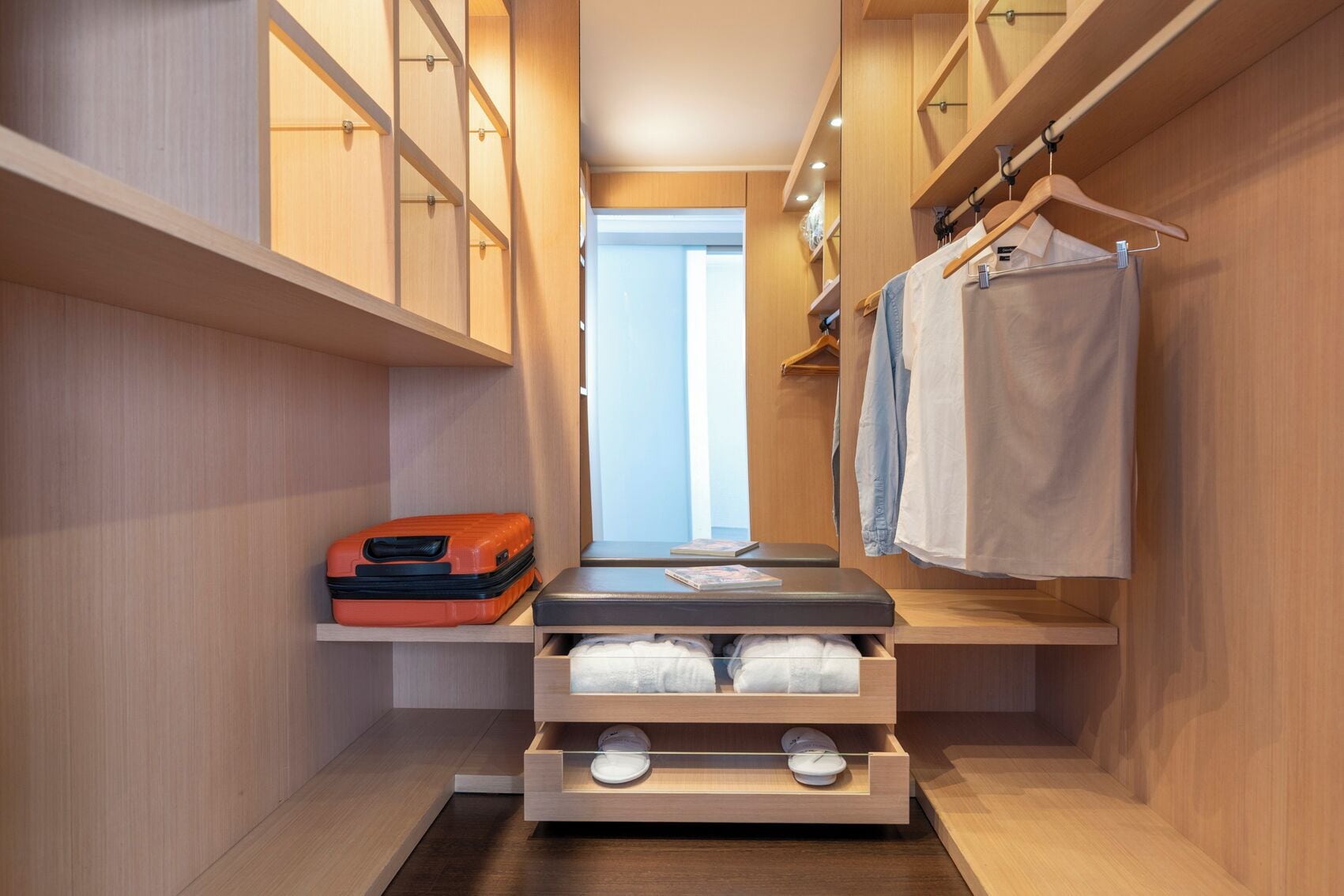 Un vestidor estrecho con estanterías de madera, un banco con cajones y ropa colgada.