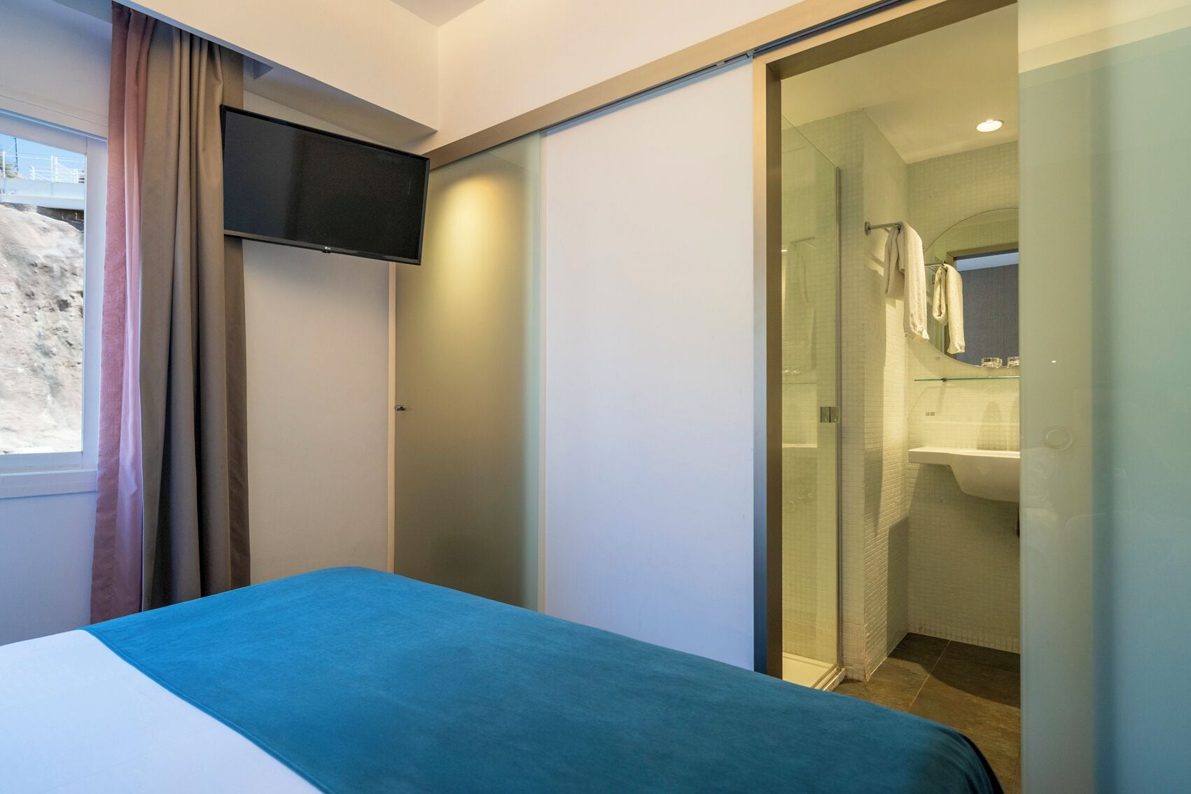A bedroom with a bed, a TV, a sliding door to a bathroom, and a view of the outside.