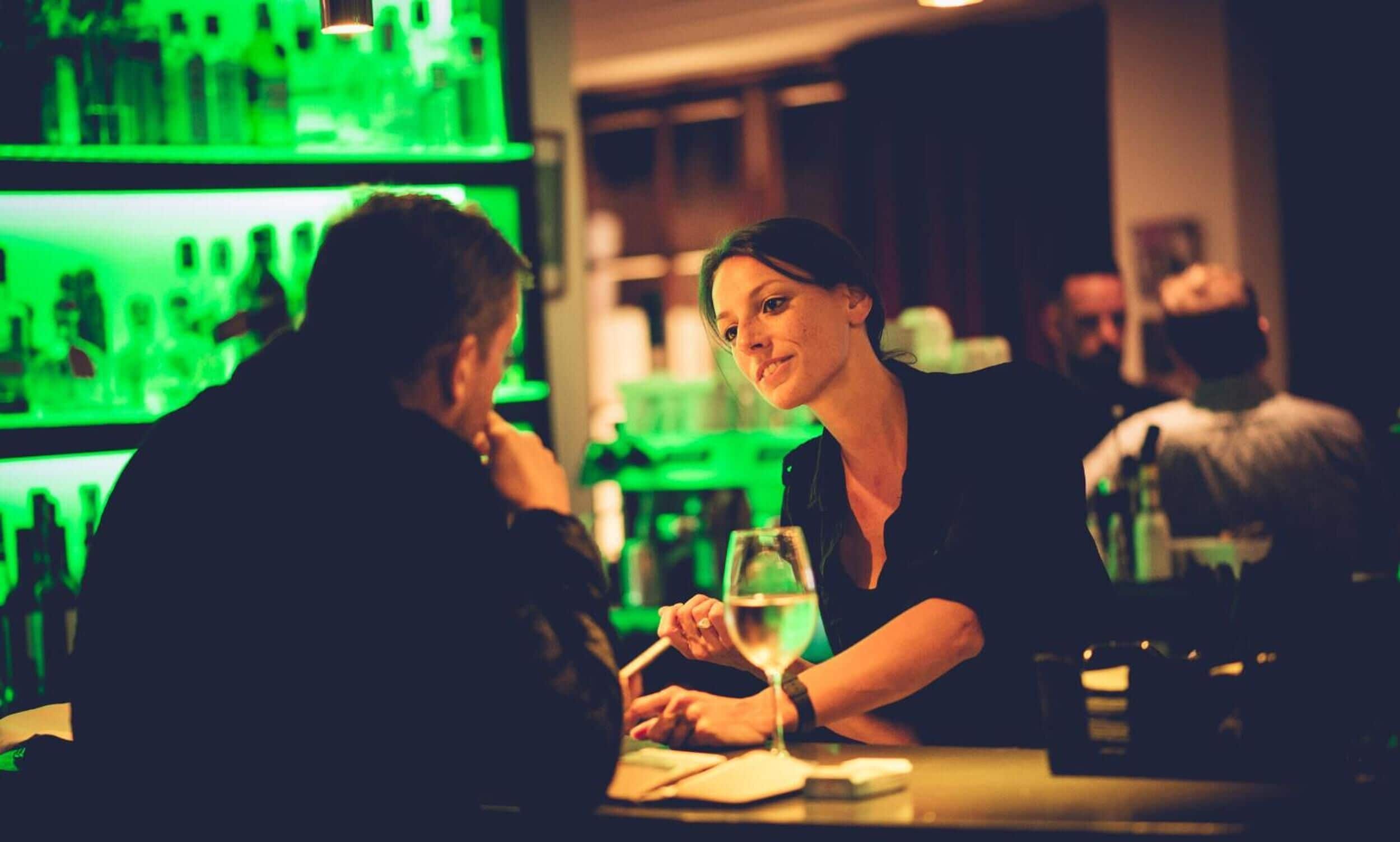 A bar setting with a green-lit background and a person holding a glass.