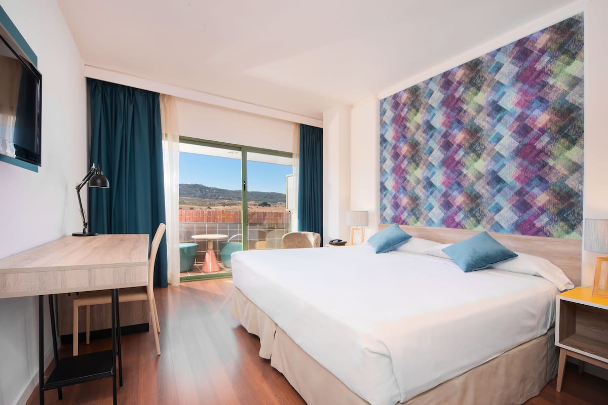 Ein Hotelzimmer mit Bett, Schreibtisch und Stuhl, einem großen Fenster mit Blick und einem farbenfrohen Wandmuster.