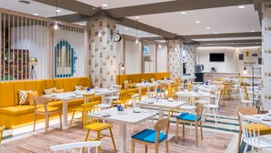 A modern restaurant with yellow and white seating, a kitchen area, and a clock on the wall.