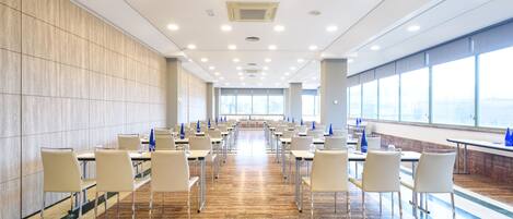 A spacious conference room with rows of tables and chairs, large windows, and a polished wooden floor.