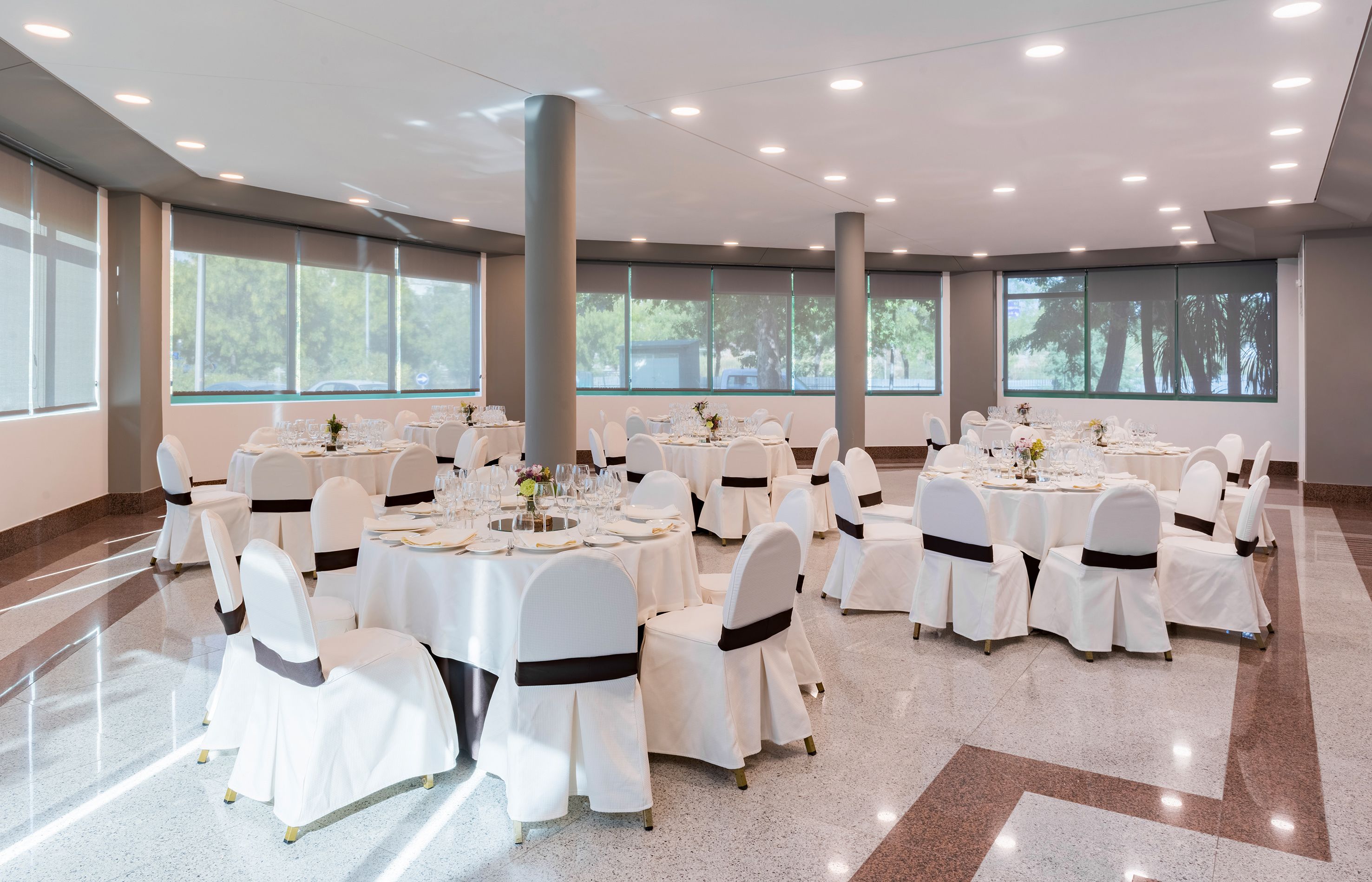Une salle de banquet spacieuse, avec des tables rondes, des chaises blanches et de grandes fenêtres.