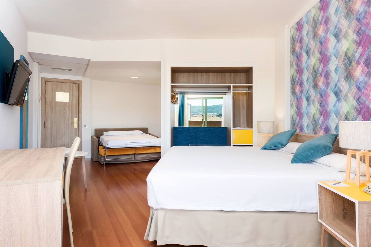 Ein Hotelzimmer mit Bett, Schreibtisch, Stuhl, Wandfernseher und einem Fenster mit Ausblick ins Freie.