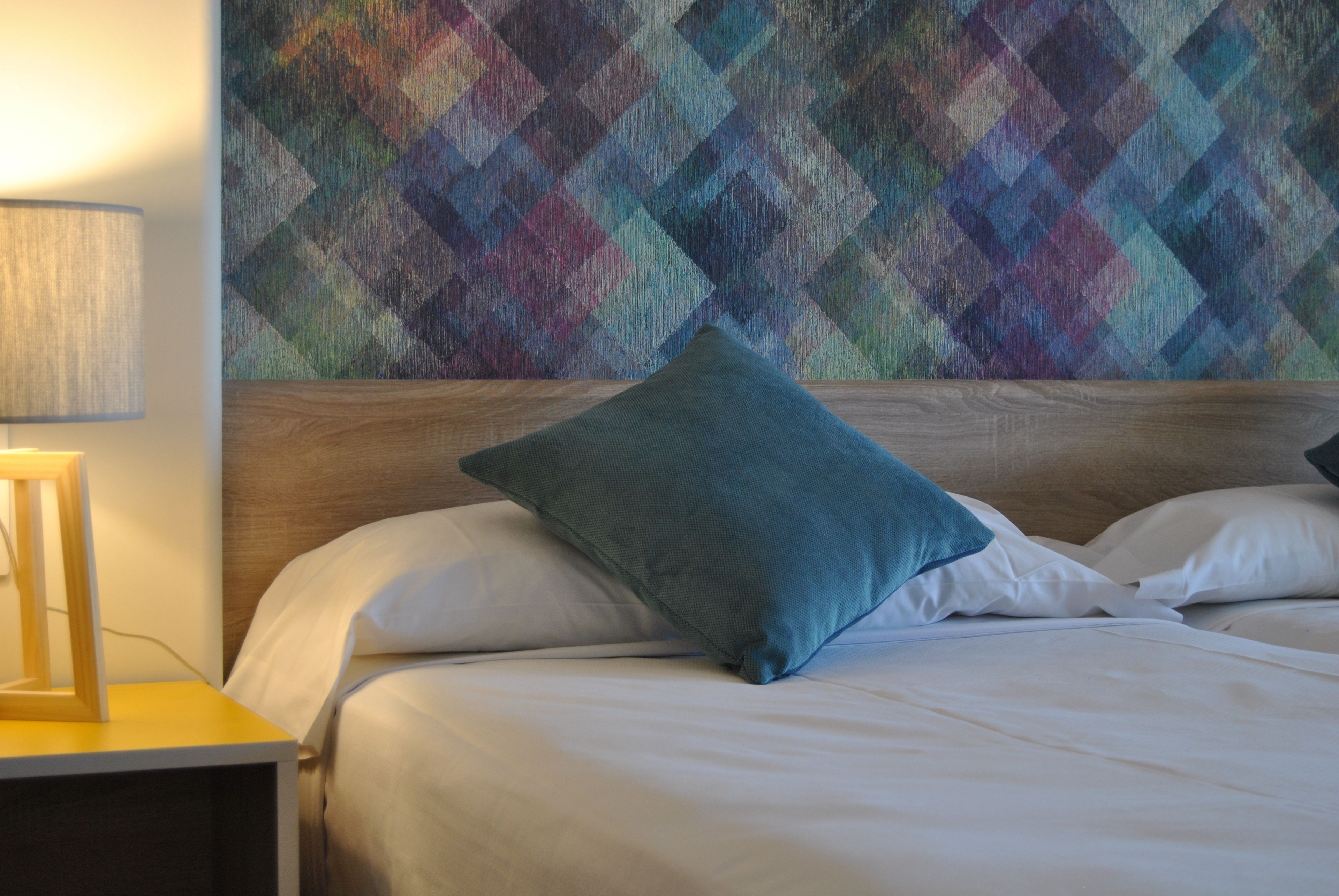 Una cama bien tendida con una almohada azul, un cabecero de madera y un tapiz de pared colorido.