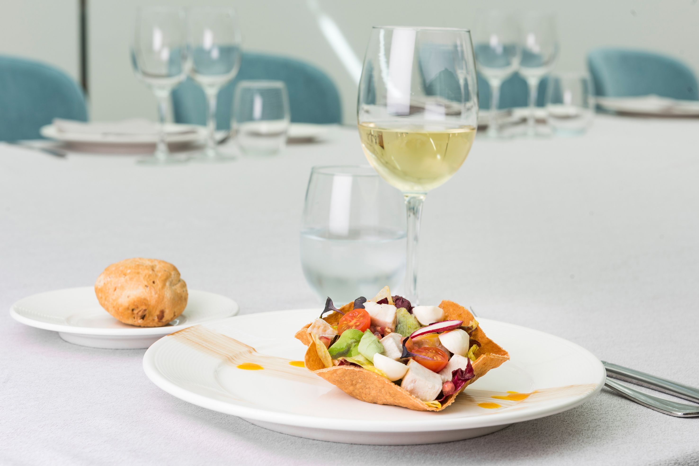 Una mesa de comedor con un plato de comida, una copa de vino blanco y una galleta.