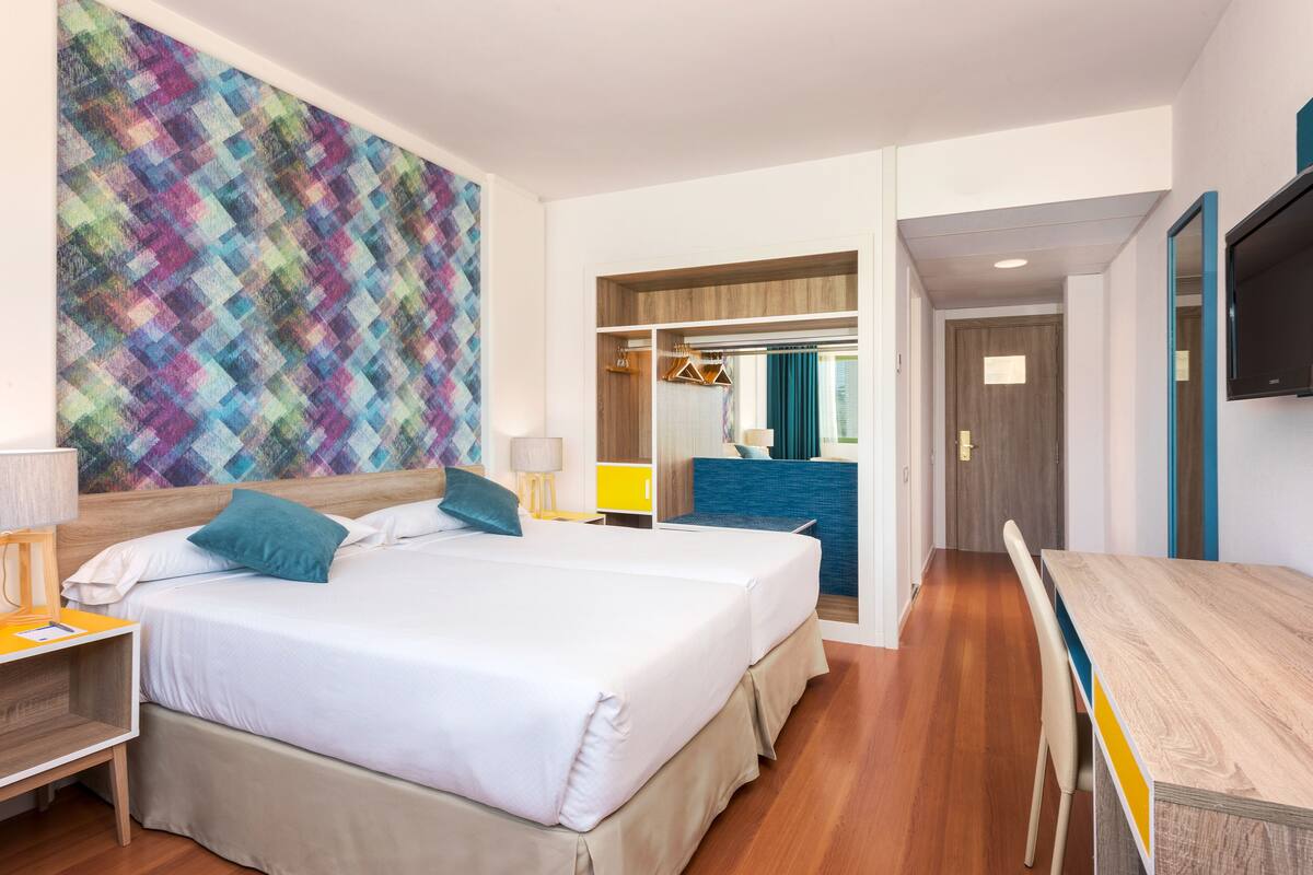 Ein Hotelzimmer mit einem großen Bett, einem Schreibtisch und einem Fernseher. An der Wand hängt ein farbenfrohes Wandbild, über dem Bett befindet sich ein Spiegel.
