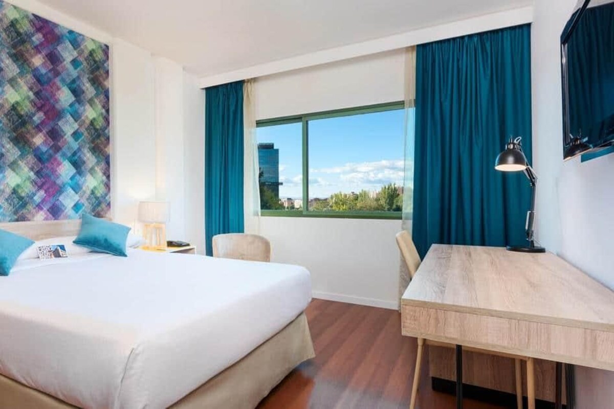 Habitación de hotel con una cama grande, un escritorio, una silla, una ventana con cortinas y una obra de arte de colores en la pared.