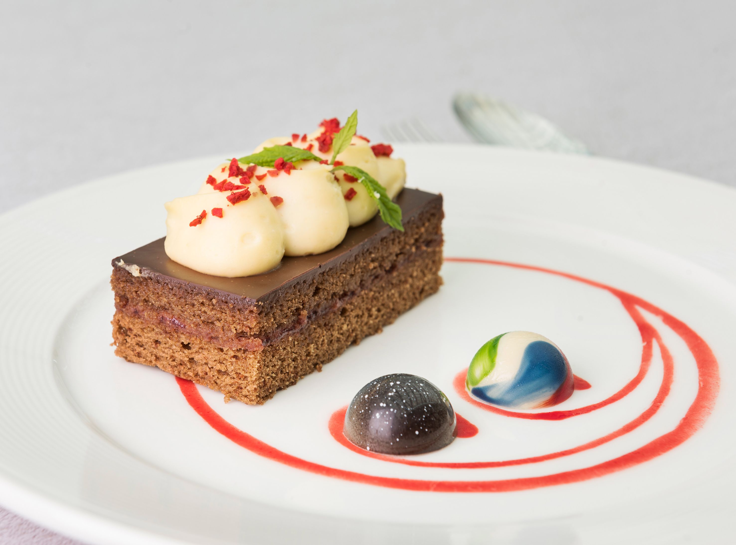 Un plato de postre con una rebanada de pastel cubierta de crema y frutos rojos, una esfera de chocolate y un postre esférico azul y blanco.