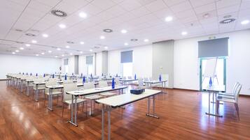 Uma sala de conferências com fileiras de mesas e cadeiras, paredes brancas e piso de madeira.