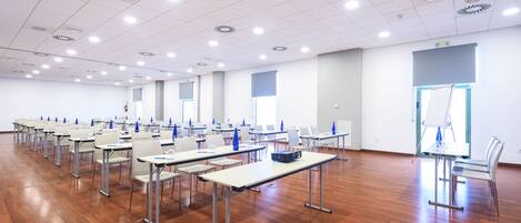 Une salle de conférence avec des rangées de tables et de chaises, des murs blancs et un plancher en bois.