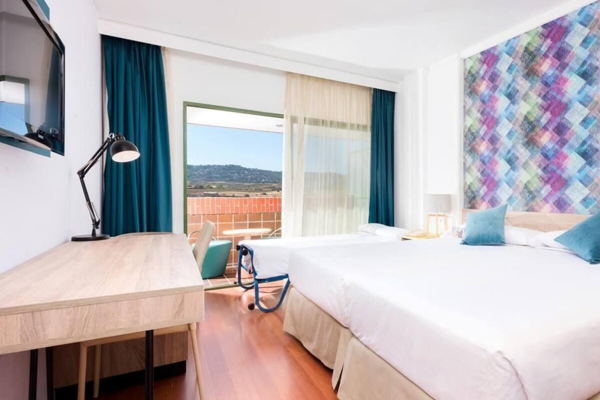 Habitación de hotel con una cama grande, un escritorio, una silla, un balcón con vistas y una pared colorida.
