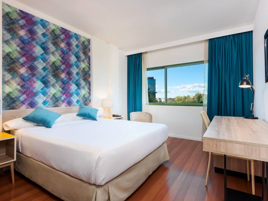 Habitación de hotel con una cama grande, un escritorio, una silla, una ventana con cortinas y un tapiz de pared colorido.