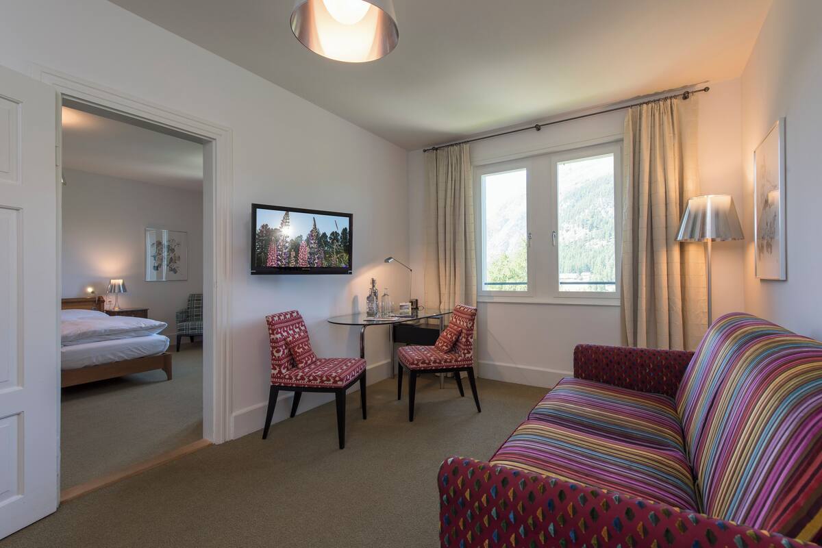 A hotel room with a bed, a desk with chairs, a colorful sofa, and a TV on the wall.