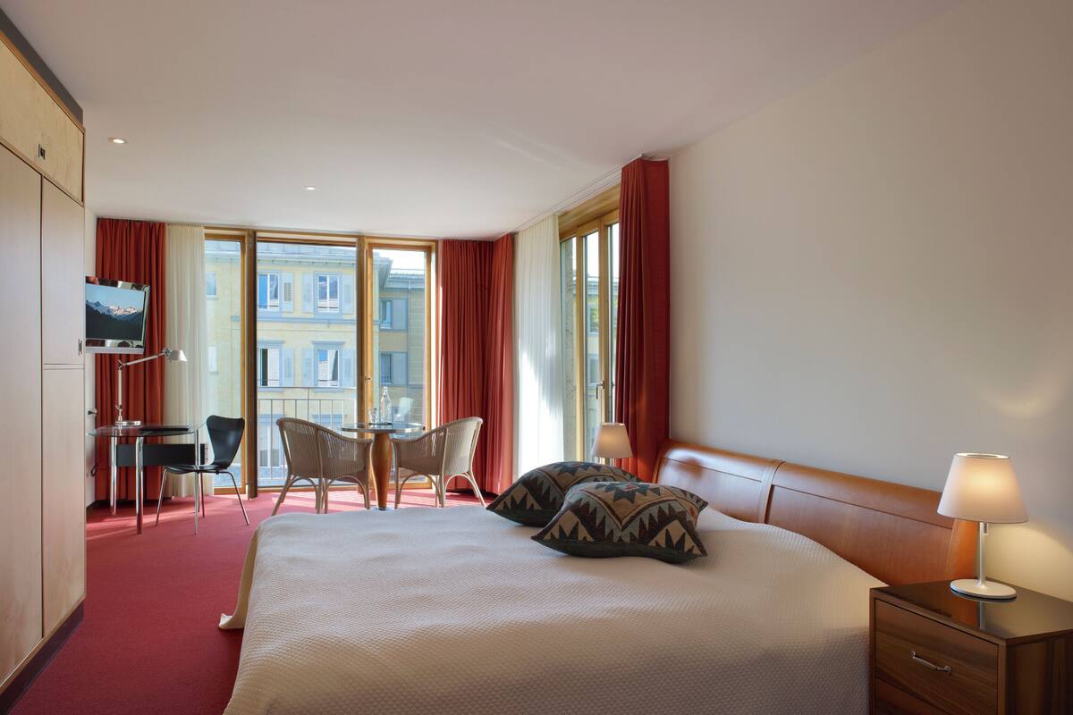 A hotel room with a large bed, a desk with two chairs, a television, and a view of buildings through large windows.