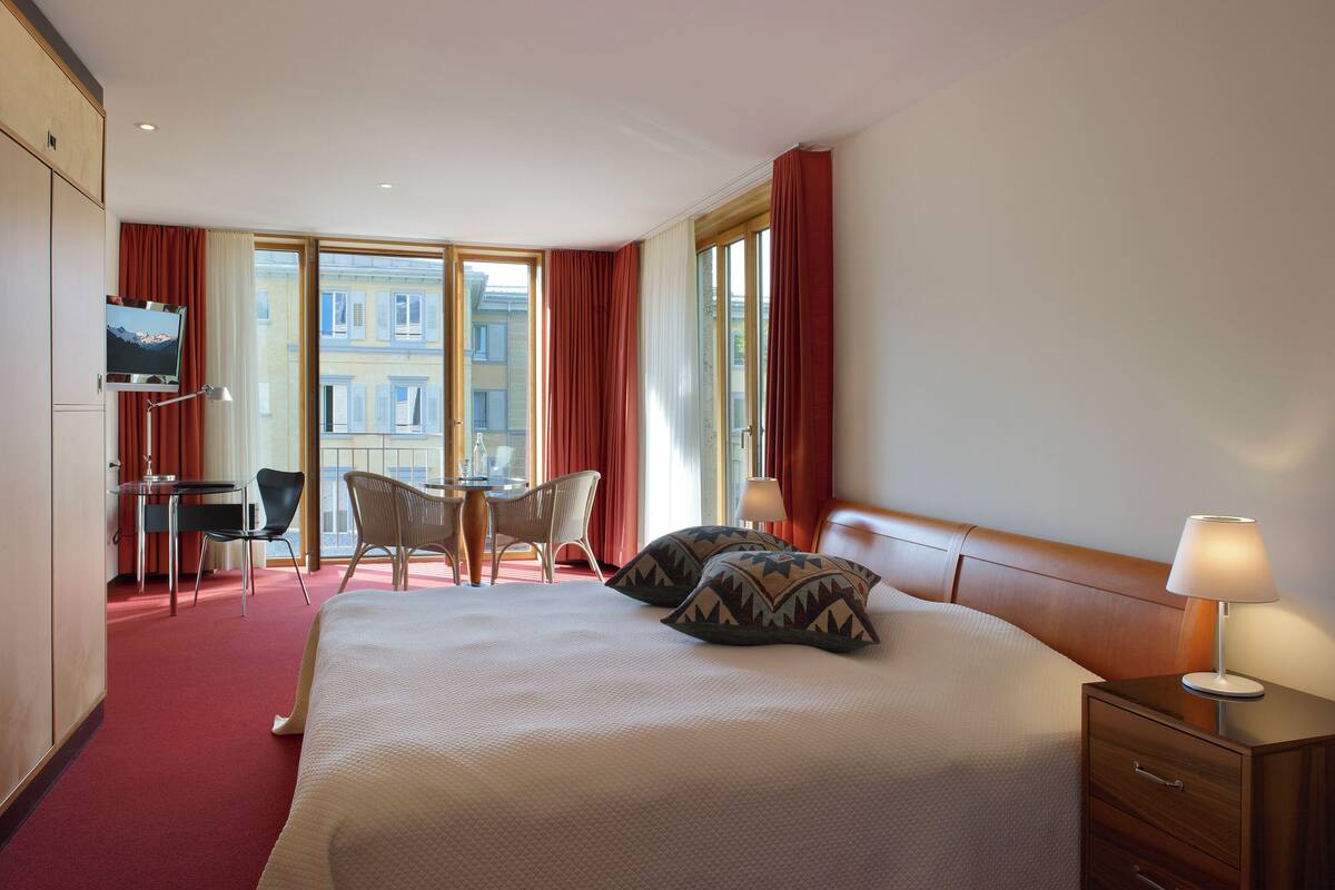 A hotel room with a bed, a desk, a chair, a TV, and large windows.