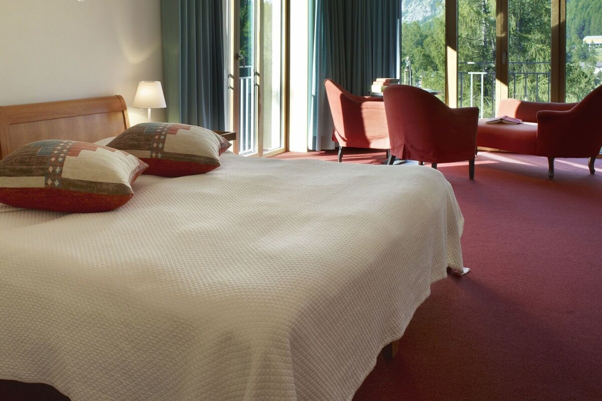 A neatly made bed with a wooden headboard, two bedside lamps, and a red chair in a room with a red carpet.