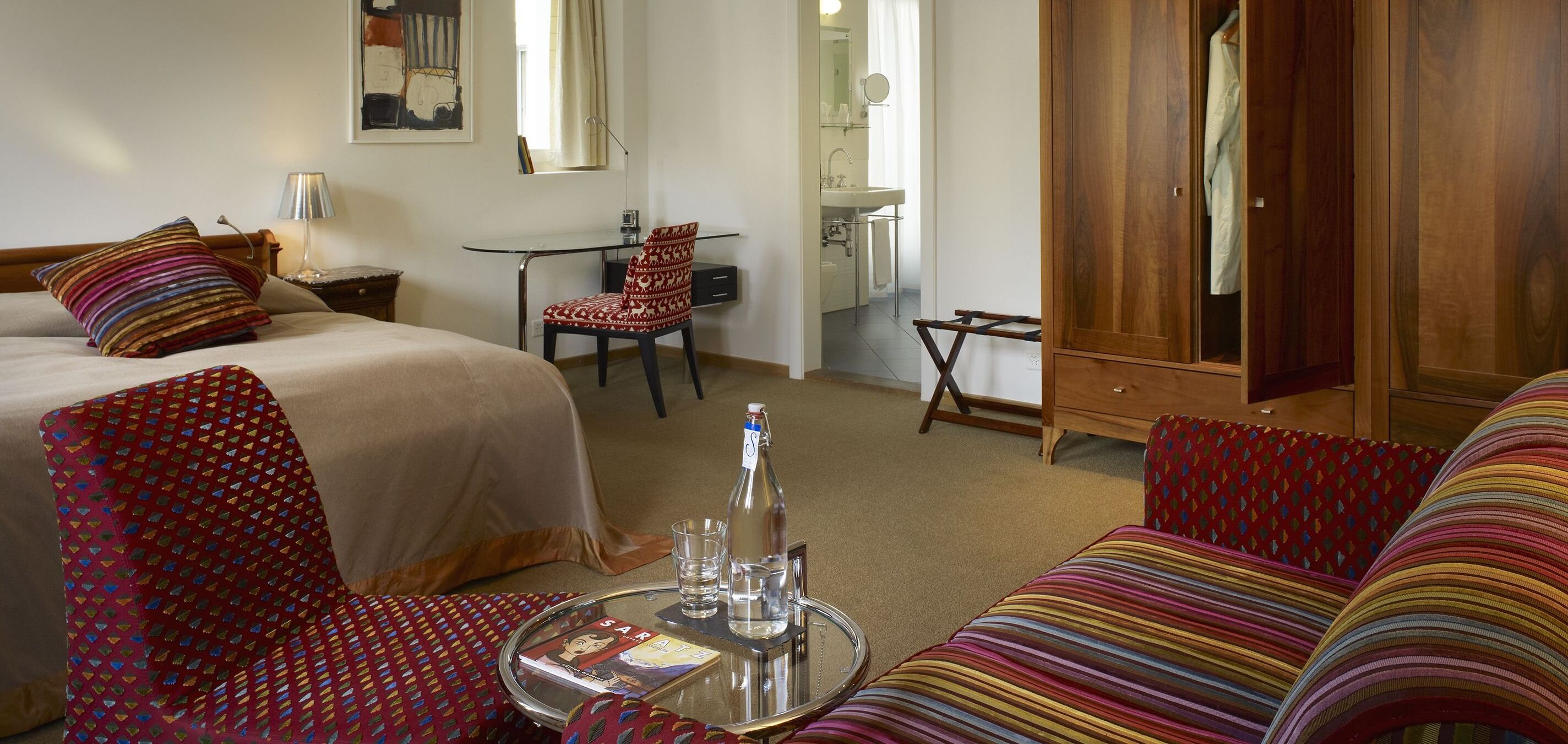 Habitación de hotel con cama, escritorio, sillón, sofá y una mesita con una botella y vasos.