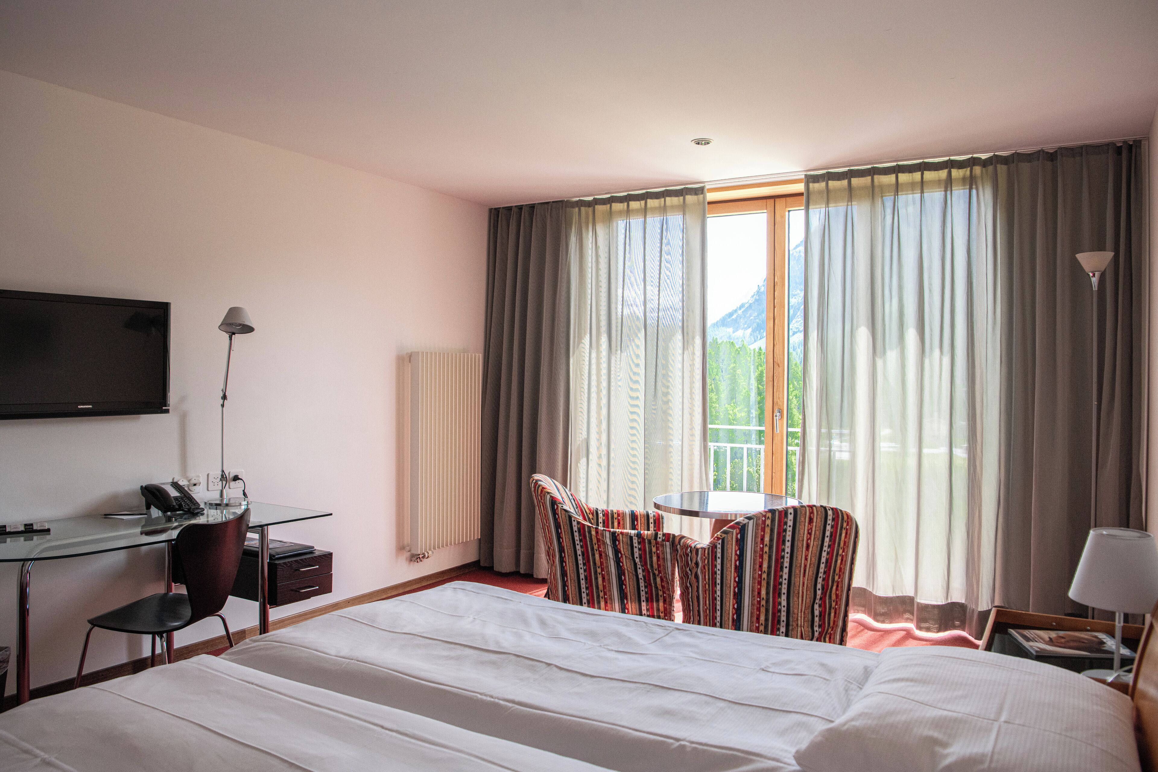 A hotel room with a bed, a desk with a chair, a TV, and a window with curtains.