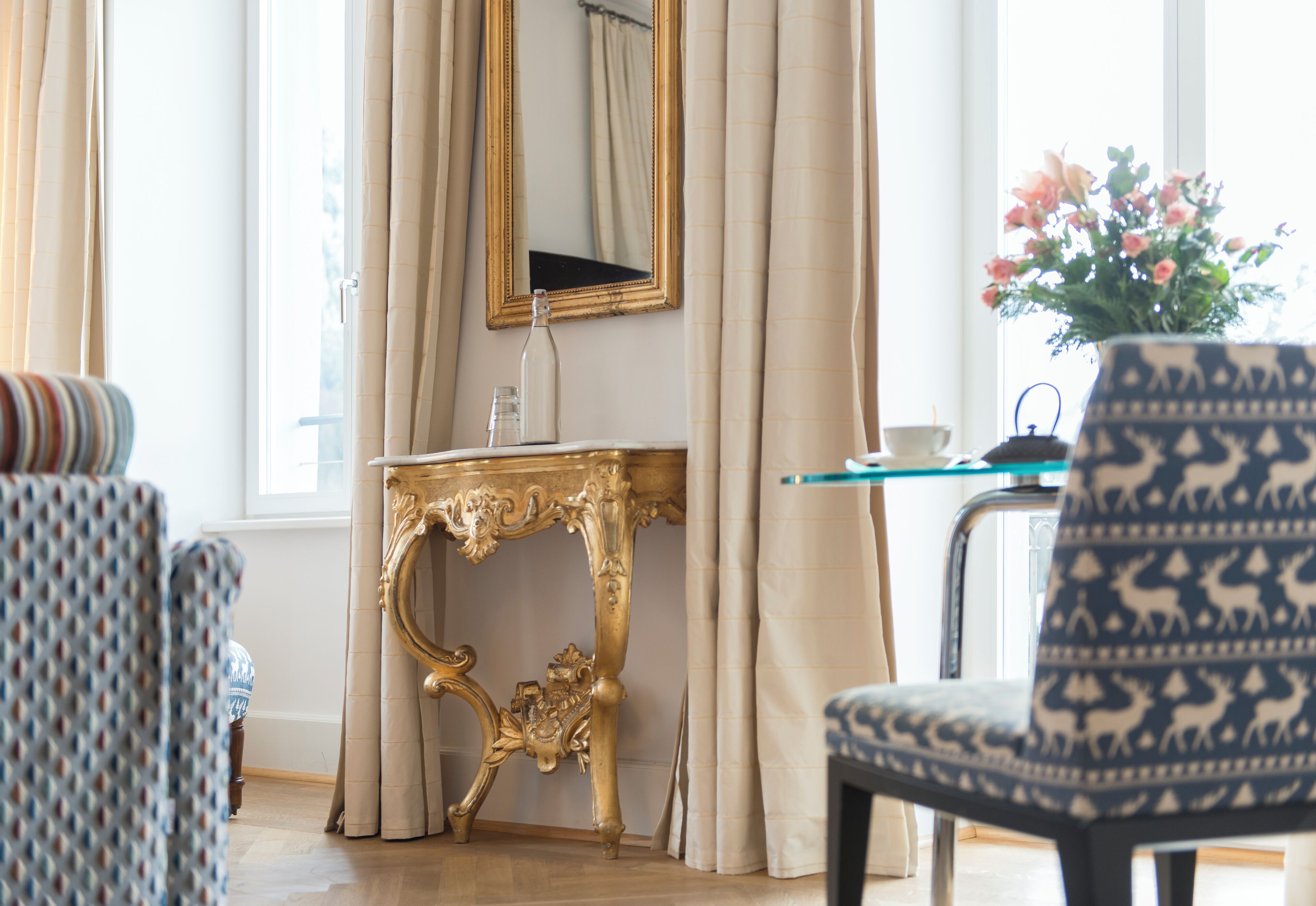 A room with a patterned armchair, a gold ornate side table, a mirror, and a vase with flowers.
