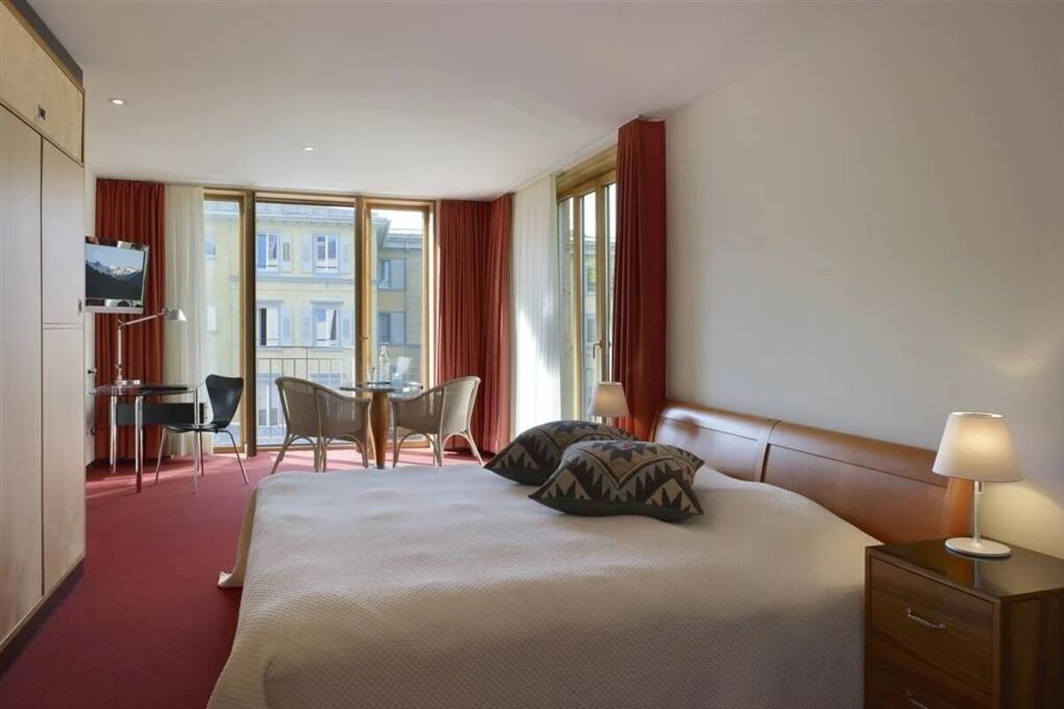 A hotel room with a bed, a desk, a chair, a TV, and large windows.