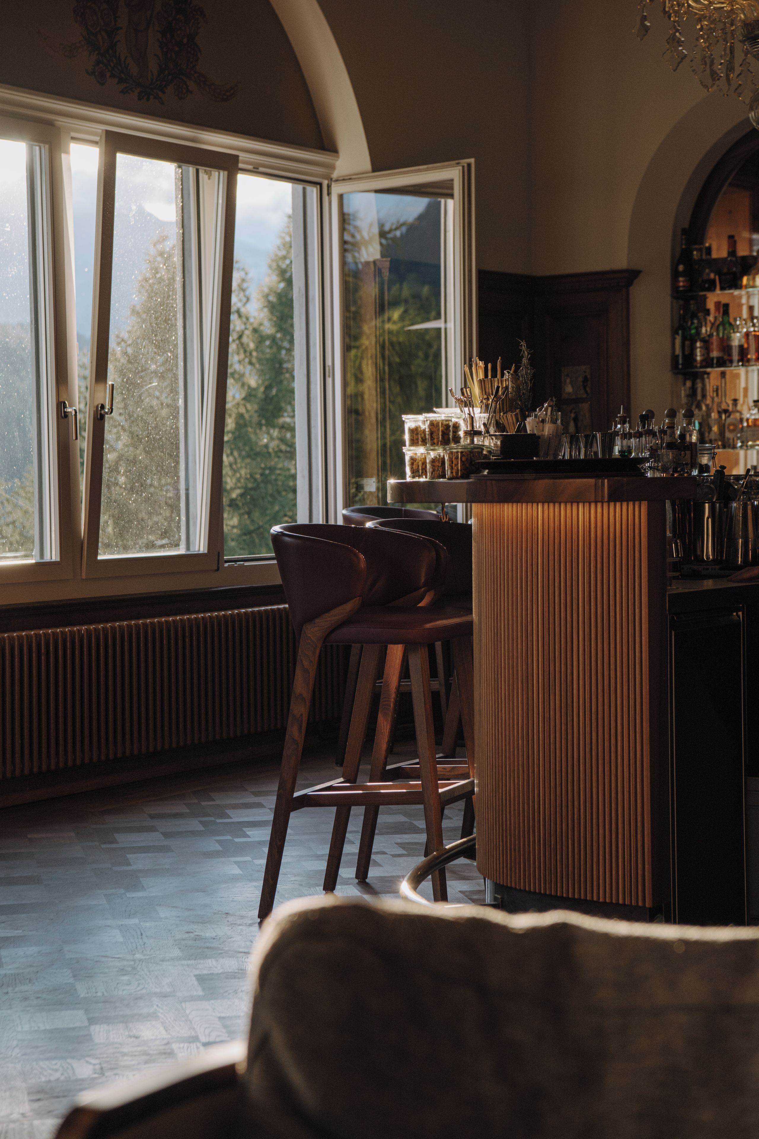 Eine Bar mit Hockstühlen, einer Theke und Blick auf Bäume durch große Fenster.